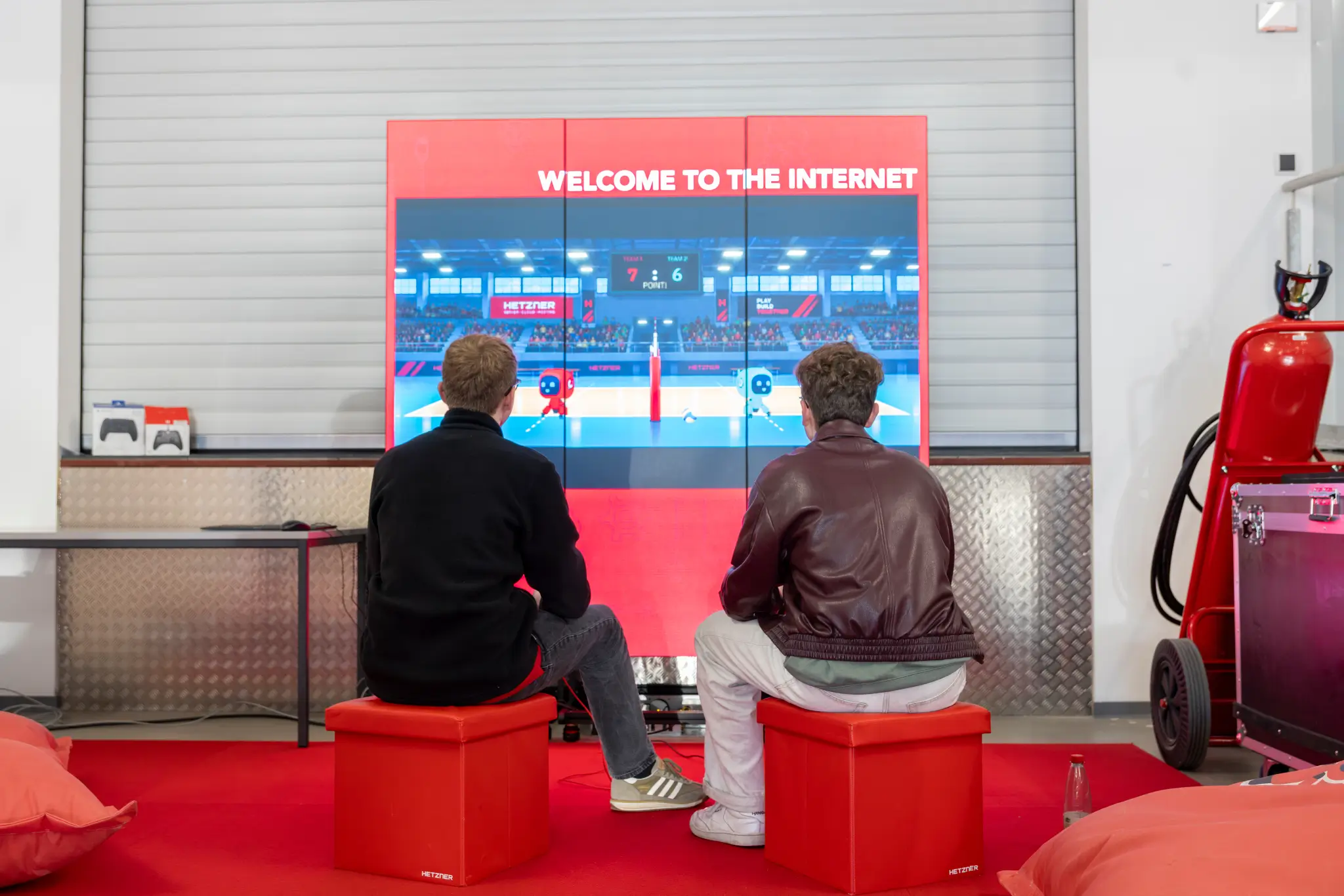 Two people sit on red stools in the gaming area.