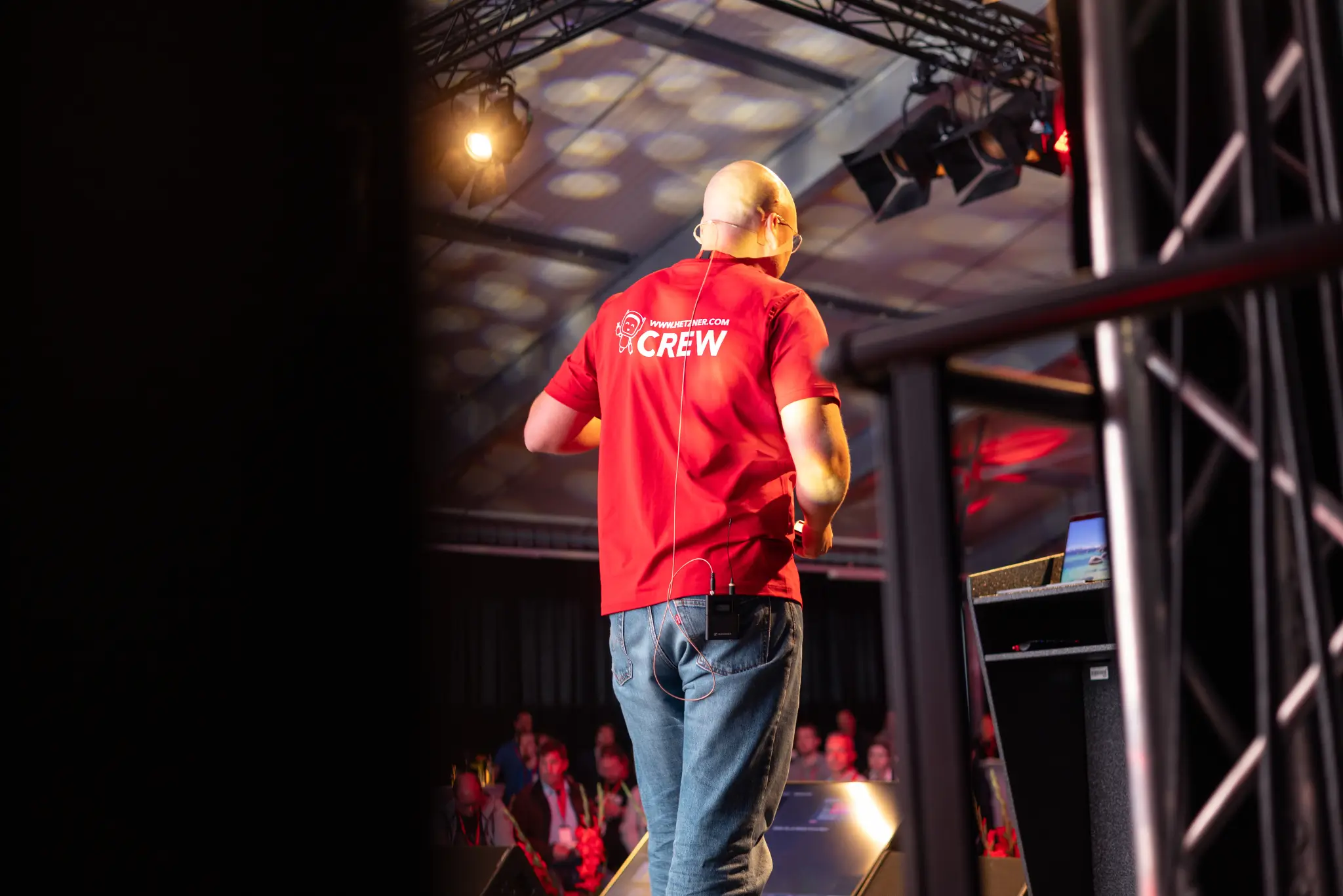Speaker in a red Hetzner Crew shirt seen from behind on a stage holding talking about digital sovereignty.
