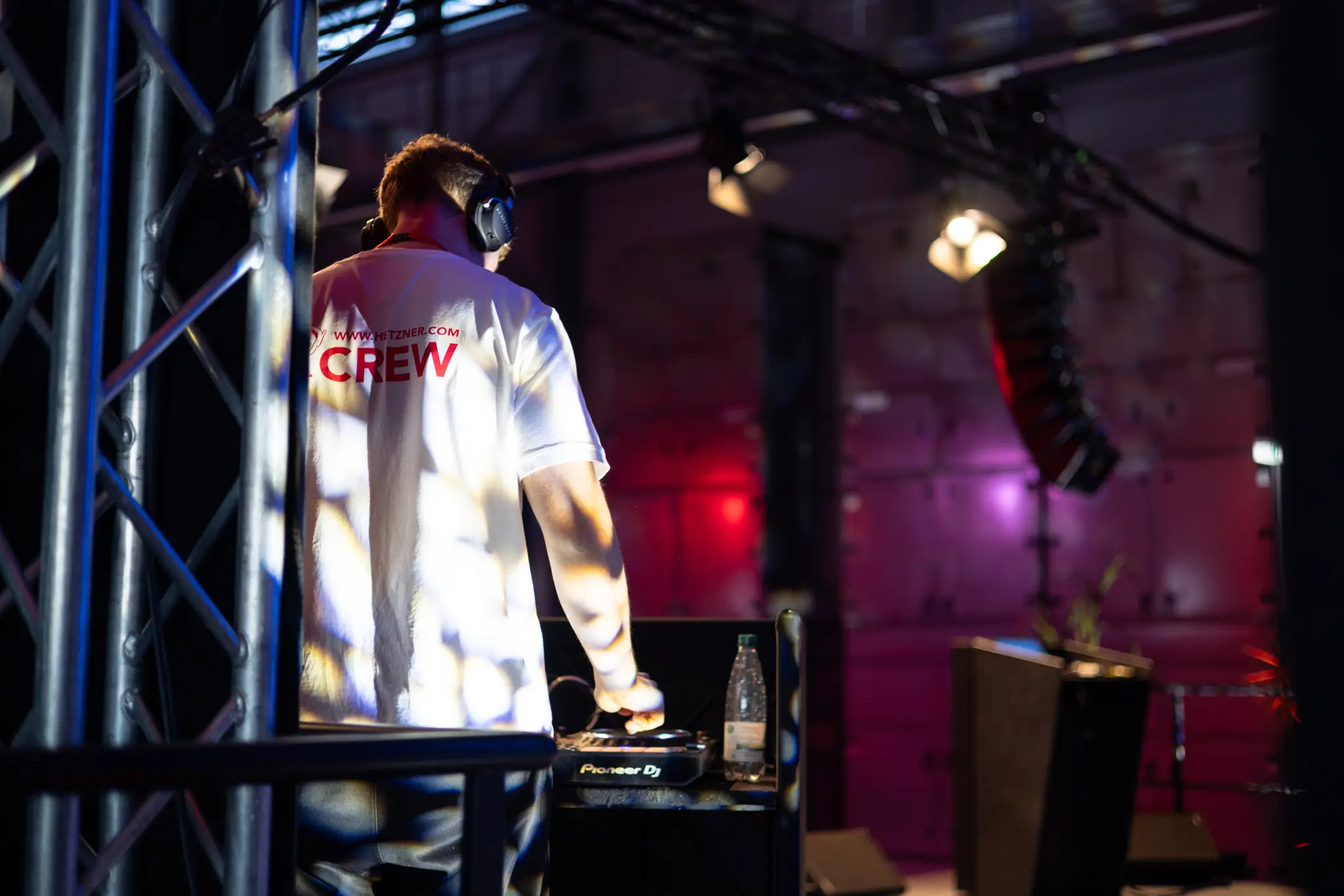 DJ in a white Hetzner Crew shirt seen from behind in front of stage equipment and spotlights.