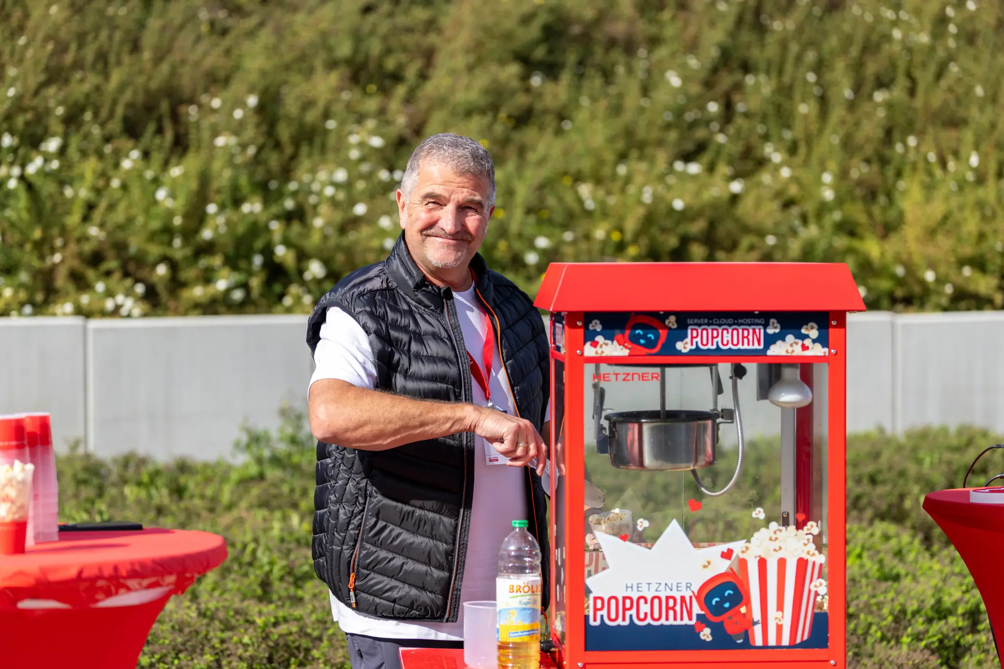 Smiling man stands next to a red Hetzner popcorn machine outdoors.