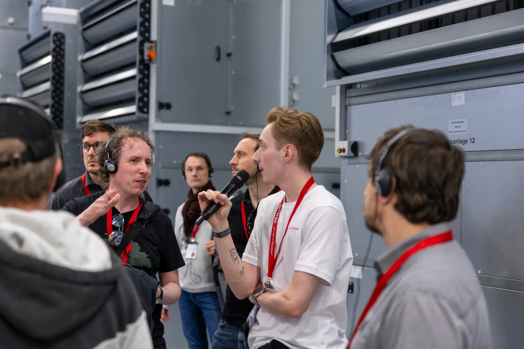 Group wearing headsets and lanyards in front of Hetzner air cooling system, Hetzner employee explaining into a microphone.