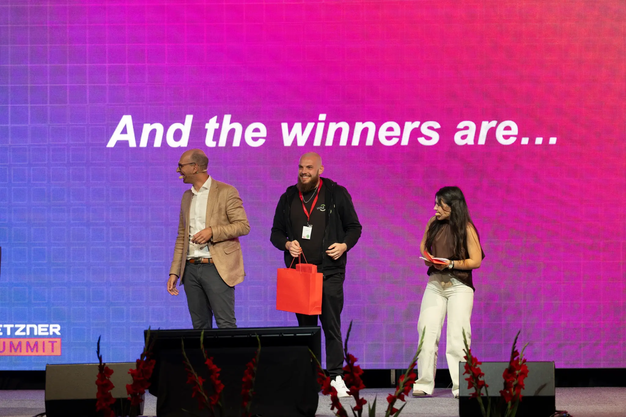 Hetzner CEO Stephan Kornikova and host standing on stage in front of a screen reading "And the winners are…" providing gifts to the contests winners.