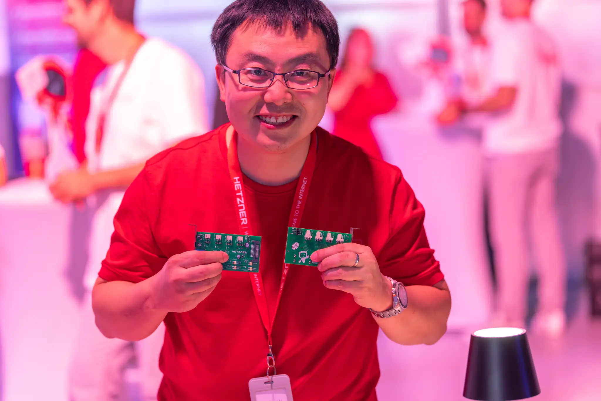 Smiling man in a red Hetzner shirt holds two green circuit boards toward the camera.